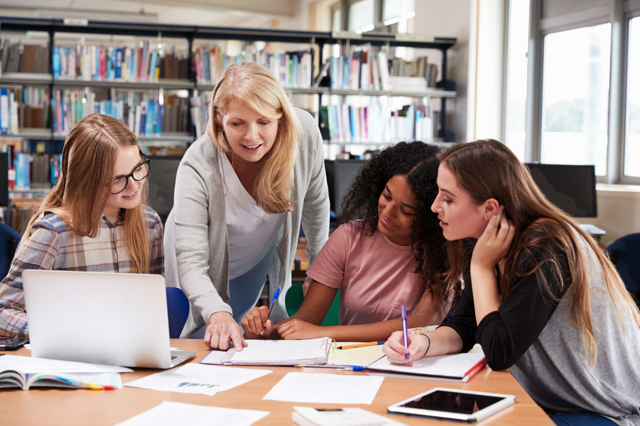 Eine Lehrerin hilft den Schülerinnen an einem Bibliothekstisch, um den herum Laptops, Notebooks und Lernmaterialien liegen. Helle, lernfördernde Umgebung.