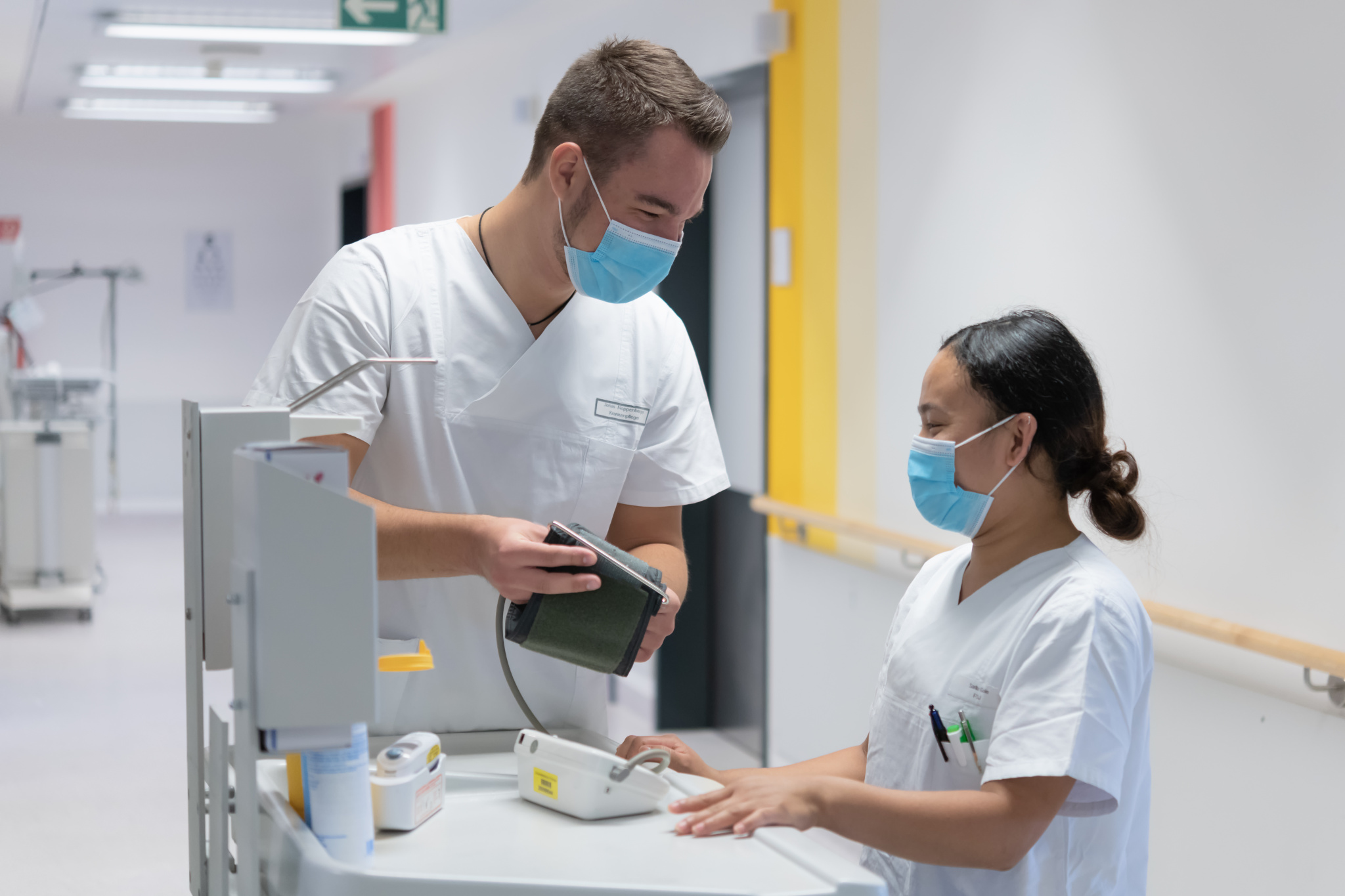 Zwei Menschen mit Kitteln und OP-Maske stehen in einem Krankenhausflur. Der eine gibt der anderen ein Blutdruck-Messgerät.