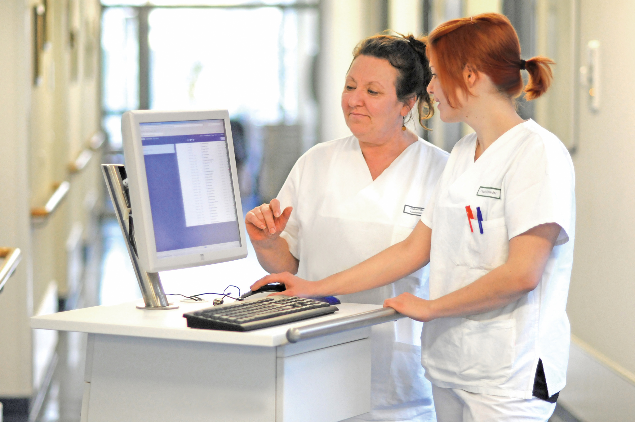 Eine FSJlerin und ihre Anleitung stehen in einem Krankenhausflur und füllen gemeinsam die Pflegedokumentation aus.