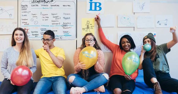 Fünf Freiwillige (FSJ/BFD) sitzen beim Seminar auf einer Bank, lachen, winken und blasen Luftballons auf.