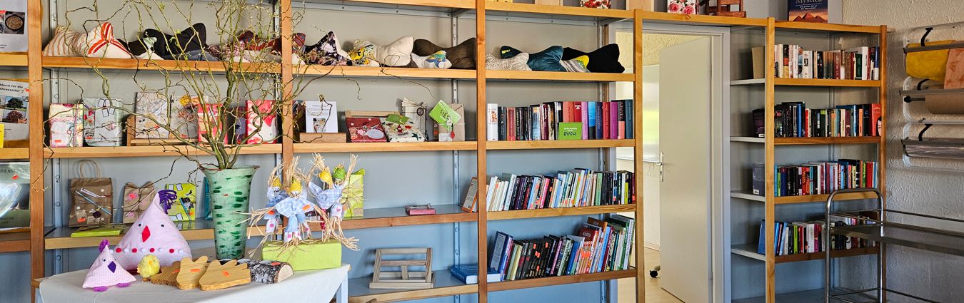 Innenraum mit großen Holzregalen, die mit Büchern, Körben und handgefertigten Dekorationsobjekten gefüllt sind. Auf einem Tisch im Vordergrund stehen Bastelarbeiten und eine Vase mit Zweigen. Rechts führt eine offene Tür in einen angrenzenden Raum, und weiteres Material ist in Regalen gelagert.