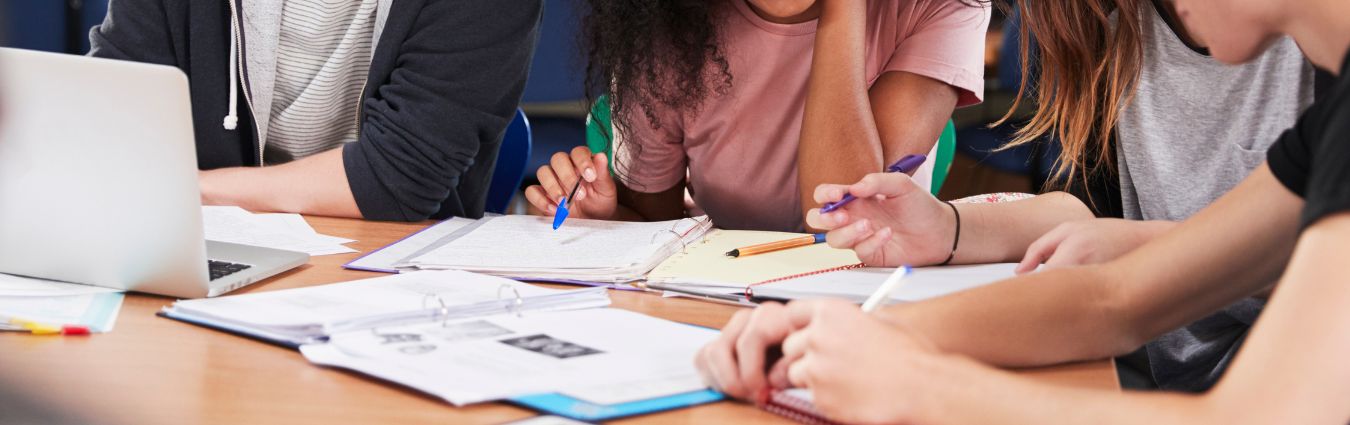 Ein Tisch mit Laptop, Heften und Papier, an dem die Arme junger Menschen zu sehen sind, die konzentriert mit Stiften arbeiten.