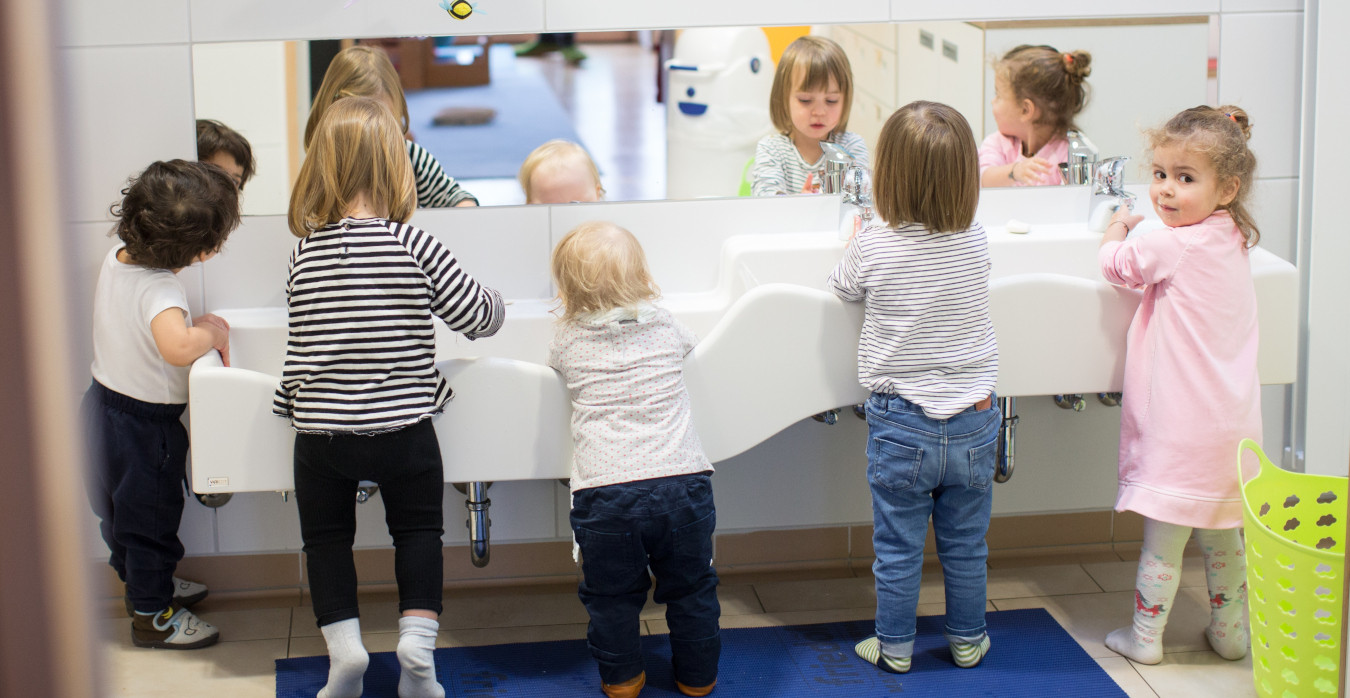 Fünf Kleinkinder stehen an einem Waschbecken vor einem Spiegel, vier sind von hinten zu sehen. Ein Kind dreht sich und blickt in die Kamera.