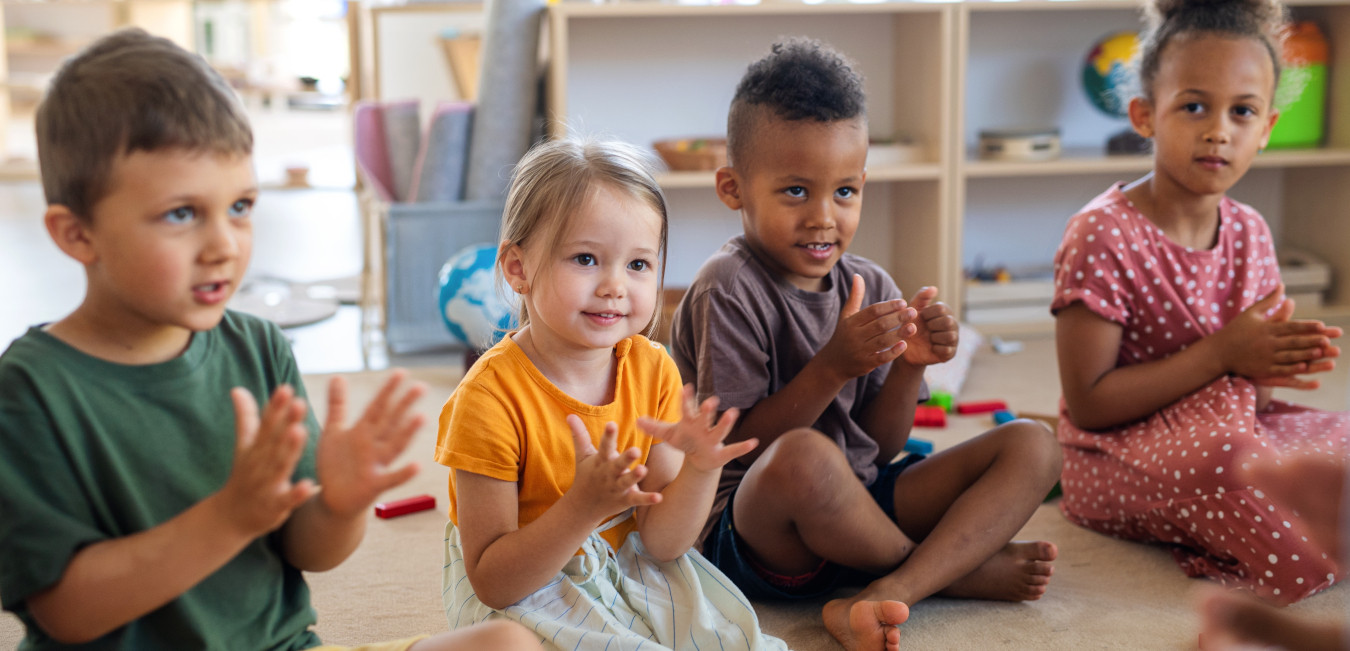 Vier Kleinkinder sitzen in einem Raum auf dem Boden und Klatschen in die Hände. Im Hintergrund sind Spielsachen zu sehen. 