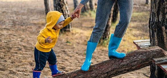 Eine Erwachsene Person hält ein ca. 2 jähriges Kind an der Hand. Beide stehen auf einem Baumstamm und tragen Gummistiefel.