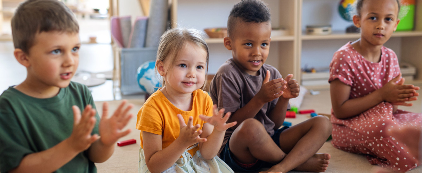 Vier Kleinkinder sitzen in einem Raum auf dem Boden und Klatschen in die Hände. Im Hintergrund sind Spielsachen zu sehen. 