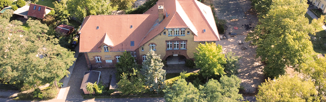 Luftaufnahme eines großen, historischen Backsteingebäudes mit rotem Ziegeldach, umgeben von vielen Bäumen und einem weitläufigen Außenbereich. Das Gebäude ist die Kita Nuthewinkel.