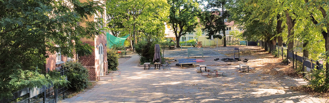 Schattiger Außenbereich mit gepflastertem Weg, Sitzgelegenheiten, Bäumen und angrenzendem Backsteingebäude; dahinter ein offenes Gelände mit Spiel- und Aufenthaltsflächen.