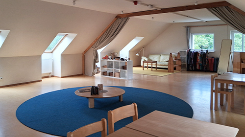 Heller Dachraum mit Holzbalken, Dachfenstern und großem blauen Teppichkreis; niedrige Regale, Sitzmöbel und ein Kleiderbereich stehen an den Wänden.