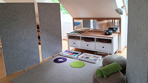Gemütliche Lese- und Spielecke unter einer Dachschräge mit Trennwänden, niedrigem Regal mit Materialien und Kisten, kleinen Teppichen, Sitzkissen und Beleuchtung; im Hintergrund ein offener Raum mit Tisch und Teppich.