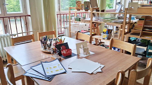 Heller Gruppenraum mit großem Holztisch und Stühlen; auf dem Tisch liegen Papier, Stifte und Bastelmaterialien, im Hintergrund stehen offene Regale mit verschiedenen Materialien und Gegenständen, große Fenster sorgen für viel Tageslicht.