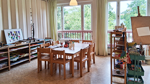 Heller Gruppenraum mit großem Tisch und kleinen Stühlen in der Mitte; auf dem Tisch stehen Becher mit Stiften, im Raum befinden sich offene Regale mit Materialien, eine Staffelei und große Fenster mit Blick ins Grüne sorgen für viel Tageslicht.