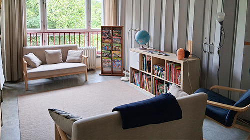 Ruhiger Raum mit zwei Sofas und einem niedrigen Tisch; an der Wand steht ein Regal mit Büchern und ein Globus, daneben eine Stehlampe, große Fenster geben den Blick ins Grüne frei.