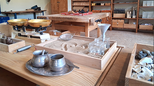 Raum mit Holzarbeitstisch und verschiedenen Materialien; im Vordergrund stehen Messingwaagen, Metallgefäße und ein Tablett mit Sand und Glasgefäßen, im Hintergrund befinden sich Regale mit Werkzeugen und Arbeitsmaterialien.