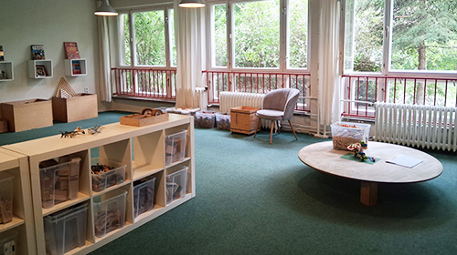 Heller Raum mit großem Fensterbereich und Blick ins Grüne; im Raum stehen ein niedriges Regal mit Materialien, ein runder Tisch mit Spielgegenständen, ein Sessel sowie weitere kleine Möbel und Boxen entlang der Wände.