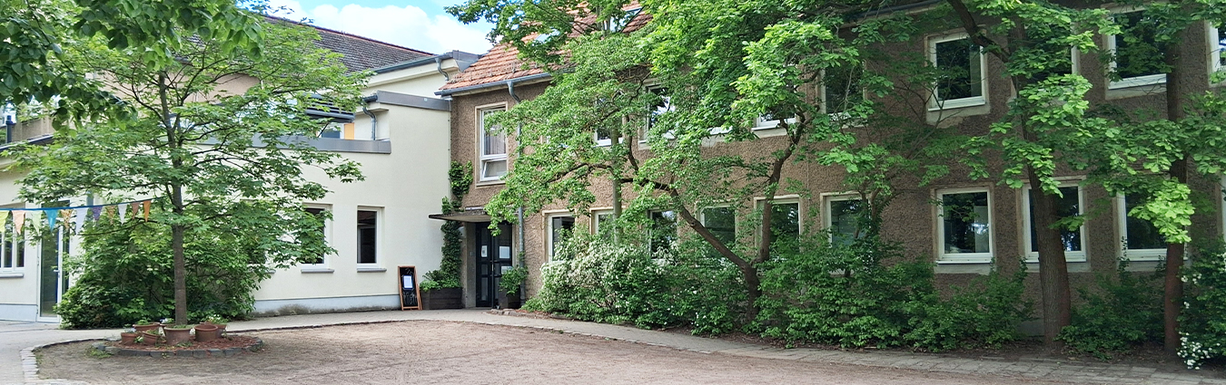 Innenhof mit begrünten Gebäuden und Bäumen, gepflasterter Fläche und dekorativen Wimpelketten, Eingang zu einem Haus im Hintergrund.