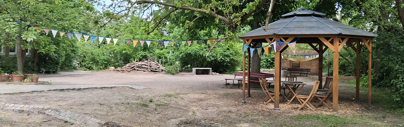 Garten mit Holzgazebo und Sitzmöbeln, umgeben von Bäumen und Girlanden, im Hintergrund offener Platz mit Feuerstelle und Holzstapeln.