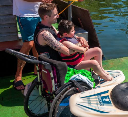Laufeingeschränkter Junge sitzt auf dem Schoß seines Pädagogen. Beide sitzen auf einem Surfboard und sind bereit um auf das Wasser gezogen zu werden. Im Vordergrund steht sein leerer Rollstuhl.