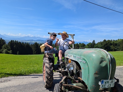 Rollstuhlfahrer und älterer Herr sitzen gemeinsam auf einem alten Traktor mit einem wunderschönen Bergpanorama im Hintergrund