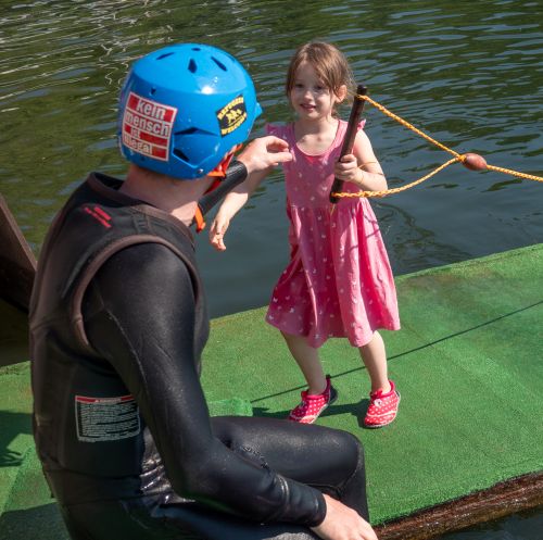 Weibliches Vorschulkind reicht seinem Pädagoge im Neoprenanzug eine Handel um mit dem Surfboard zu starten. Auf dem Helm des Pädagogen steht groß geschreiben "Kein Mensch ist illegal"