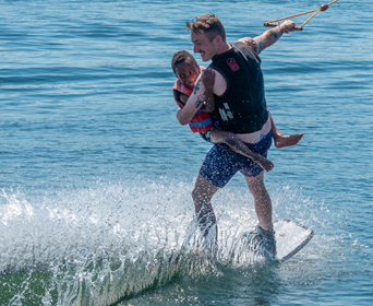 Kleiner Junge auf dem Arm seines Pädagogen. Beide surfen mit dem Wakeboard übers Wasser