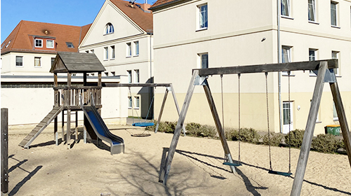 Spielplatz mit Kletterturm aus Holz, Rutsche und Schaukelgestell auf sandigem Untergrund vor einem hellen, mehrstöckigen Gebäude mit rotem Dach.