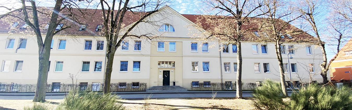 Breites, zweistöckiges helles Gebäude mit rotem Satteldach, zahlreichen Fenstern und einem zentralen Eingang; im Vordergrund stehen mehrere kahle Bäume und Büsche unter blauem Himmel.