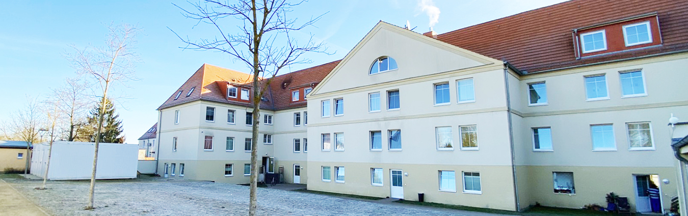 Helles, mehrstöckiges Gebäude mit rotem Satteldach und zahlreichen Fenstern; im Vordergrund ein gepflasterter Hof mit einzelnen kahlen Bäumen unter klarem Himmel.