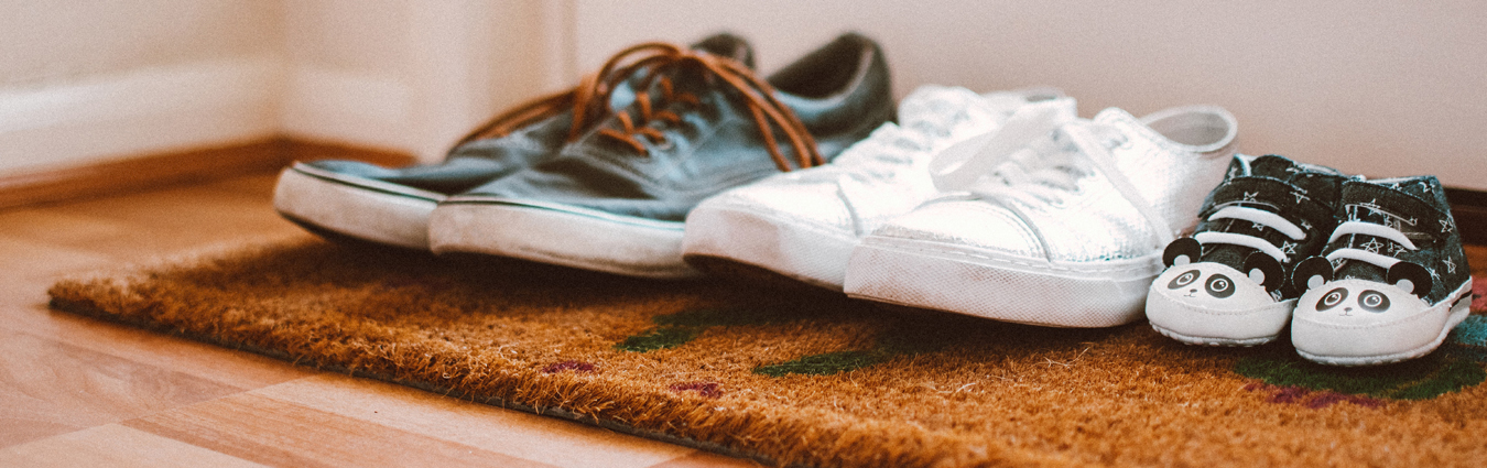 Mehrere Paar Schuhe – zwei Paar Erwachsenenschuhe und ein Paar Babyschuhe mit Panda-Motiv – stehen auf einer Fußmatte im Eingangsbereich.