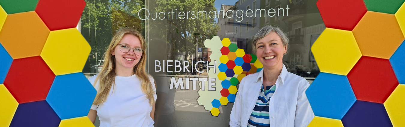 Zwei Frauen stehen lachend vor einem Schaufenster auf dem steht "Quartiersmanagement Wiesbaden". 