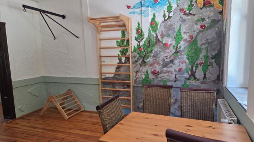 Ein Zimmer mit einem Tisch und vier Stühlen und einer Kletterwand. Die Wand ist bunt bemalt mit einem Berg und Bäumen.