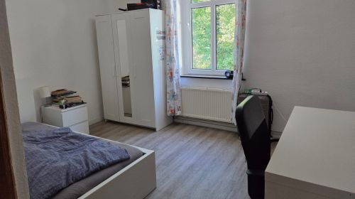 Ein Zimmer mit einem Schreitisch, einem Schreibtischstuhl, einem Fenster mit Vorhängen, einem Kleiderschrank mit Spiegel, einem Nachttisch und einem Bett. 