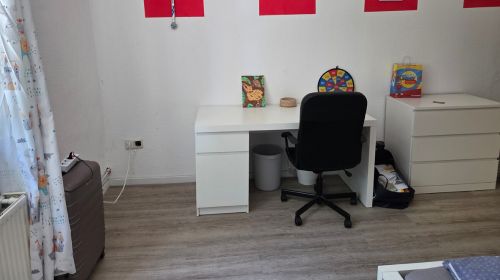 Ein Zimmer mit Schreibtisch, Schreibtischstuhl, einer Kommode und vielen roten Bildern an der Wand, ein Koffer steht in der Ecke.
