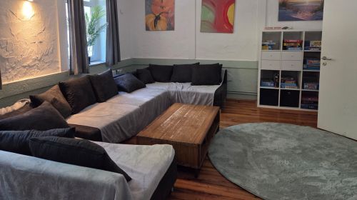 Ein Wohnzimmer mit einem großen Sofa, einem Teppich und einem Regal. An der Wand hängen einige Bilder.