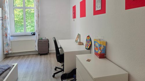 Ein Zimmer mit einem Schreibtisch mit Schreibtischstuhl und einer Kommode. Es hängen mehrere rote Bilder an der Wand.