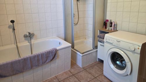 Ein Bad mit Badewanne, Dusche, Waschmaschine und einem kleinen Schrank mit Handtüchern und Kosmetik- und Hygieneartikeln.