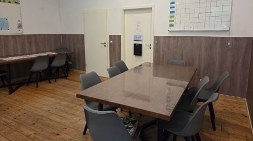 Ein Raum mit Holzboden, einem Tisch mit 8 Stühlen, und einem anderen Tisch in der Ecke mit weiteren Stühlen. An der Wand hängt ein großer Kalender