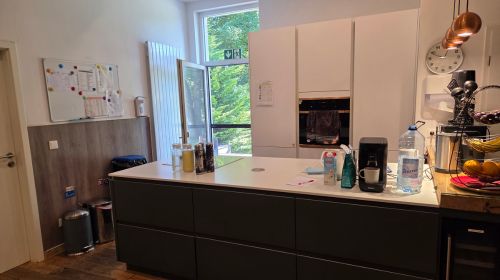 Eine Küche mit einer Kücheninsel, eine, großen Küchenschrank mit Ofen und einer Glastür, die nach draußen führt. Auf der Kücheninsel steht eine Kaffeemaschine.