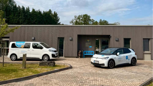 Zwei IB Autos, Dienstwagen - ein Bus und ein PKW stehen vor einem Haus mit Flachdach. 