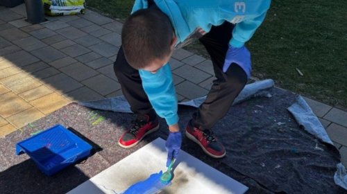 Ein Mann mit blauem Pulli ist kreativ tätig und bringt blaue Farbe auf eine Leinwand auf, die vor ihm auf dem Boden liegt. 