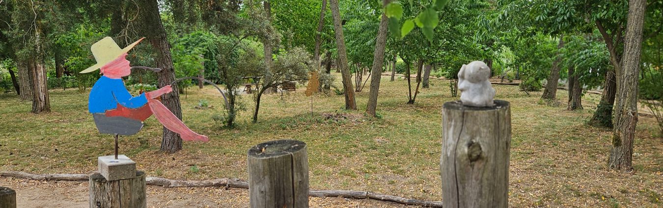 Naturnaher Garten- oder Parkbereich mit vielen Bäumen und trockenem Grasboden. Im Vordergrund stehen mehrere Holzpfosten; auf einem Pfosten ist eine bunte, dekorative Metallfigur mit Hut zu sehen, die eine Gießkanne hält. Auf einem anderen Pfosten sitzt eine kleine, helle Skulptur. Im Hintergrund erstreckt sich ein locker bewachsener, grüner Bereich mit Sträuchern und weiteren Bäumen.