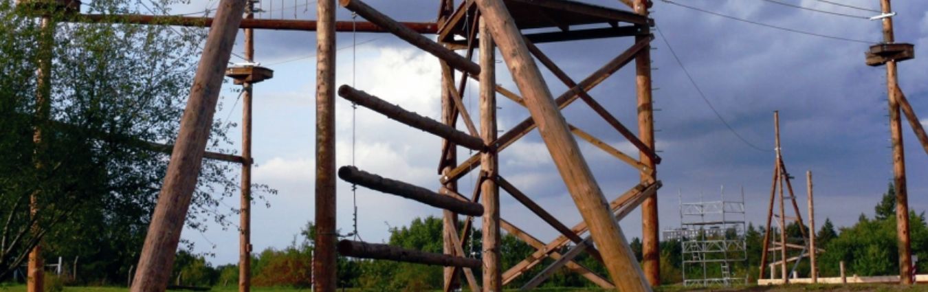 Kletter- und Balancierparcours aus Holz in einem Outdoor-Bereich. Mehrere hohe Holzbalken sind zu einer stabilen Konstruktion verbunden, zwischen denen horizontale und schwingende Elemente wie Balken und Plattformen angebracht sind. Im Hintergrund sind weitere Kletterelemente sowie eine freie, grasbewachsene Fläche mit Bäumen und ein teilweise bewölkter Himmel zu sehen.