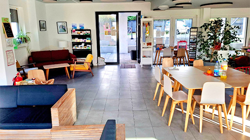 Heller, freundlich eingerichteter Gemeinschaftsraum mit Sofas, Regalen, mehreren Tischen und Stühlen; große Fenster und Pflanzen sorgen für eine einladende Atmosphäre.