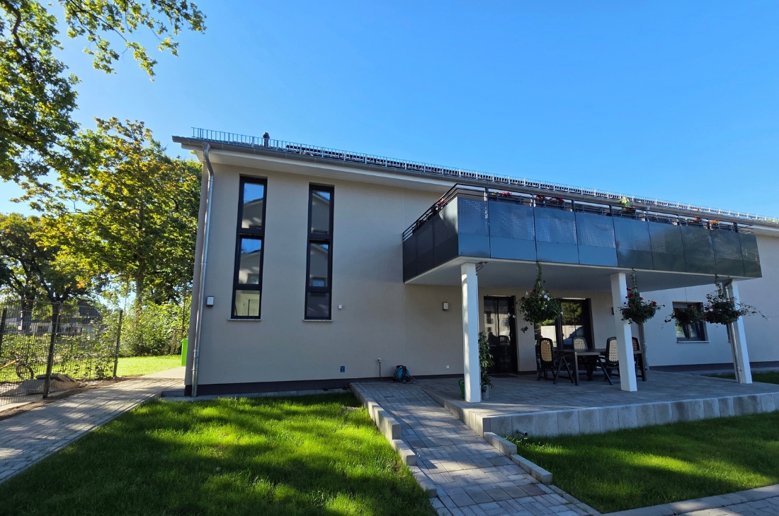 Zweigeschossiges Wohnhaus mit beiger Putzfassade und hohen, schmalen Fenstern. Im Obergeschoss ragt ein Balkon mit dunkler Lochblech-Brüstung über eine gepflasterte Terrasse mit weißen Stützen und hängenden Blumenampeln. Davor liegt ein gepflegter Rasen unter blauem Himmel.