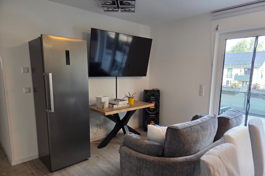 Sitzbereich mit grauem Sofa, einem an der Wand montierten Fernseher und einem Holztisch mit schwarzem X-Gestell. Daneben stehen eine große Lautsprecherbox und ein Kühlschrank aus Edelstahl. Rechts eine Balkontür mit Blick auf die Nachbarbebauung.