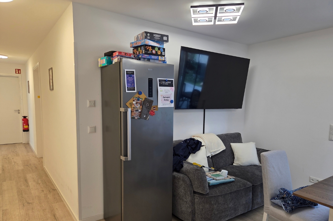 Blick vom Flur in den Wohnbereich mit grauem Sofa, Fernseher und Kühlschrank mit Aushängen und Spielen obenauf. Auf dem Sofa liegen eine Jacke, Decken und Kissen, im Vordergrund rechts ein Stuhl und der Esstisch. Im Hintergrund links ein Feuerlöscher im Flur.