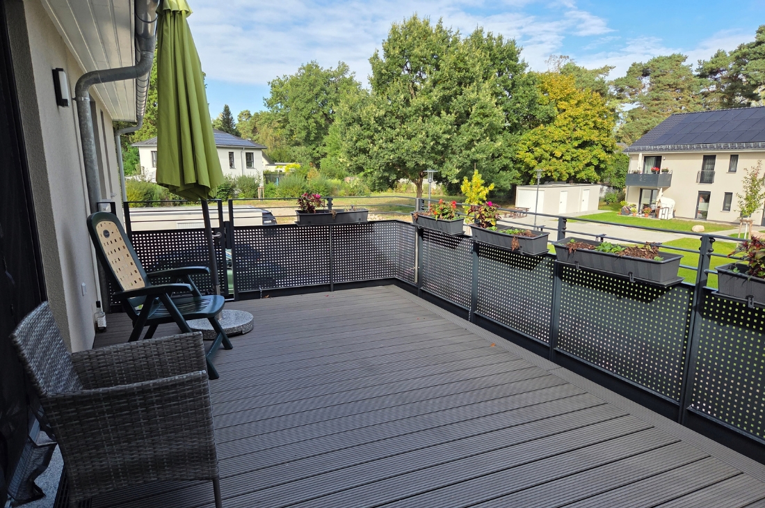 Balkon mit dunklem Dielenboden und Lochblech-Brüstung, an der Blumenkästen mit bunten Pflanzen hängen. Links stehen zwei Sitzmöbel und ein zusammengeklappter grüner Sonnenschirm. Im Hintergrund Bäume und Wohnhäuser mit Solardächern.