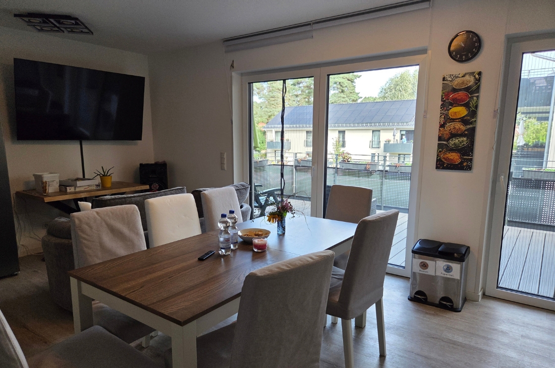 Esstisch mit Holzplatte und hellen Stühlen im Wohnbereich, auf dem Tisch Wasserflaschen, eine Schale und eine kleine Vase mit Blumen. Durch die Glasschiebetür sieht man den Balkon, an der Wand hängen eine Uhr und ein Bild mit Gewürzen. Rechts steht ein Mülltrennsystem, links Fernseher und Sofa.