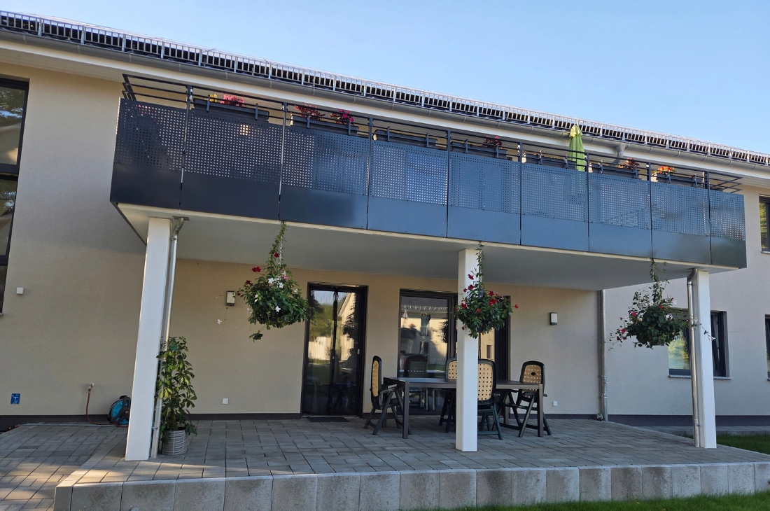 Frontansicht der überdachten Terrasse im Erdgeschoss mit Tisch und vier dunklen Kunststoffstühlen. Darüber der Balkon mit perforierter anthrazitfarbener Brüstung, Blumenkästen und einem zusammengeklappten grünen Sonnenschirm. Zwischen den weißen Stützen hängen bepflanzte Blumenampeln.