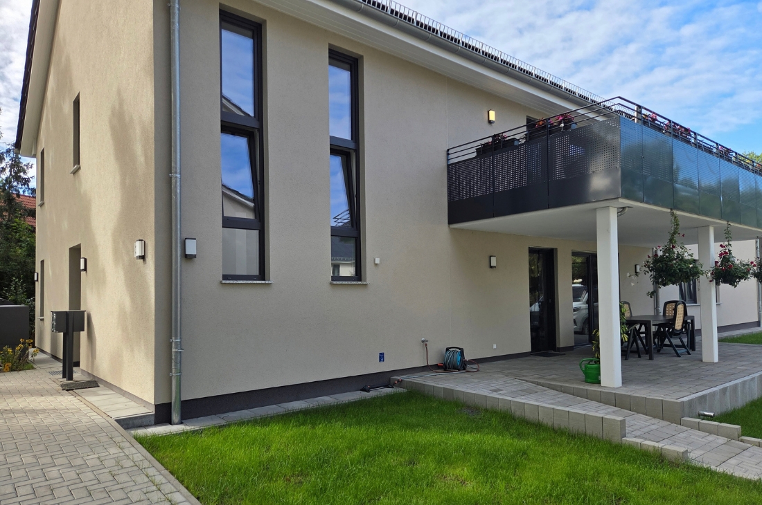 Seitenansicht des Gebäudes mit zwei hohen, schmalen Fenstern, einem Fallrohr und Wandleuchten. Rechts der Balkon mit Lochblech-Brüstung über der Terrasse, links ein Pflasterweg mit Briefkastenanlage. Im Vordergrund ein Rasenstück, am Himmel stehen leichte Wolken.